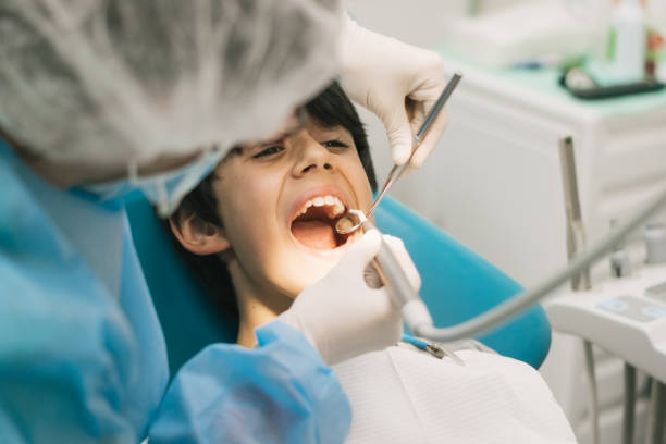 niño de pelo oscuro sentado en la silla del dentista con la boca abierta mientras el dentista explora su boca con instrumentos en el fondo - medical exam dental hygiene caucasian mask fotografías e imágenes de stock