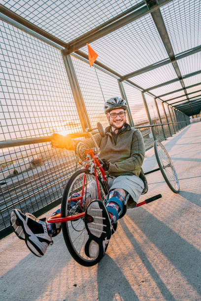 istock Handsome Young Man Smiles and Poses for the Camera While Riding a Red Handcycle Wearing Orthotic Leg Braces for Adaptive Exercise Outdoors on a Pedestrian Bridge in an Urban Sunset Setting with Copy Space 1314422804