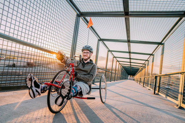 istock Handsome Young Man Smiles and Poses for the Camera While Riding a Red Handcycle Wearing Orthotic Leg Braces for Adaptive Exercise Outdoors on a Pedestrian Bridge in an Urban Sunset Setting with Copy Space 1314422808