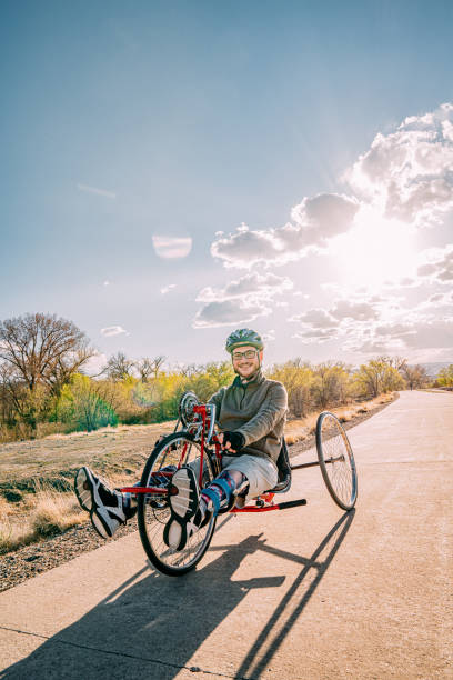 Handsome Young Man with Myelomeningocele Spina Bifida Riding a Red Handcycle Wearing Orthotic Leg Braces for Adaptive Exercise Outdoors in the Spring. Overcoming the Stigma and Challenges of Disability can be Fun on a Hand bike! amplia toma de ángulo bajo de un joven guapo sonriendo y mirando a la cámara mientras estaba sentado en su ciclo de mano rojo usando aparatos ortopédicos para las piernas para el ejercicio adaptativo al aire libre al atardecer con espacio de copia