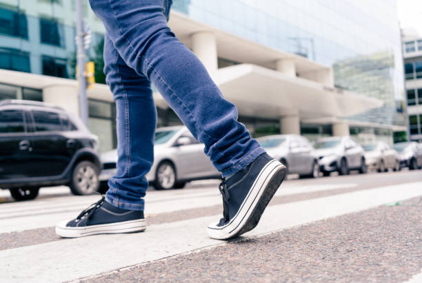 nahaufnahme der füße eines mannes in schwarzen turnschuhen, der eine straße am zebrastreifen oder fußgängerweg überquert. fläschliche bildung - pedestrian stock-fotos und bilder