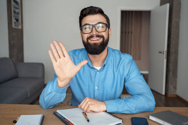 Smiling millennial caucasian man at office desk looking at camera waving hand stock photo