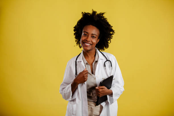 istock Attractive young female doctor in white lab coat holding digital tablet and smiling while standing against yellow background. 1314458607