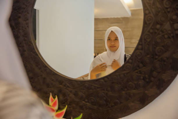 Young Indonesian Muslim Woman Fixing her Hijab on the Mirror Shot of a young Indonesian muslim woman adjusting her hijab in front of the mirror before going out for Eid Ul-Fitr salat. introspection in ramadan stock pictures, royalty-free photos & images