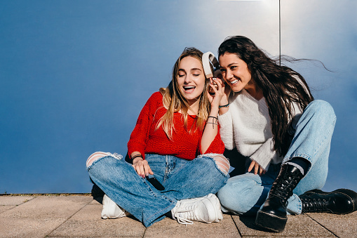 Couple of friends with long hair, one blonde and one brunette, listening to music on their cell phones sitting on the floor in front of a blue wall.