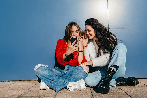 Couple of friends with long hair, one blonde and one brunette, listening to music on their cell phones sitting on the floor in front of a blue wall.