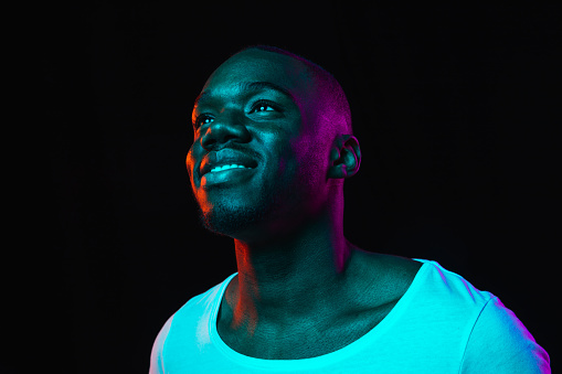 Half-length portrait of young happy joyful African-American man isolated on dark background in multicolored neon light. Concept of human emotion, facial expression, youth culture. Copy space for ad