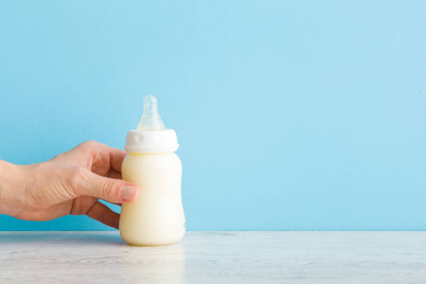 赤ちゃんの授乳のために白いミルクのペットボトルを取る若い大人の母親の手。薄い青色の壁の背景上のテキストの空の場所。パステルカラー。クローズ アップ。フロントビュー。 - 牛乳ビン ストックフォトと画像