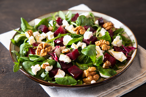 Beet or beetroot salad with fresh arugula, soft cheese and walnuts on plate, dressing and spices on dark wooden background, copy space, top view/