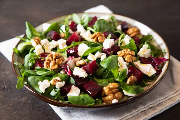 Photo of Beet or beetroot salad with fresh arugula, soft cheese and walnuts on plate, dressing and spices on dark wooden background, copy space, top view