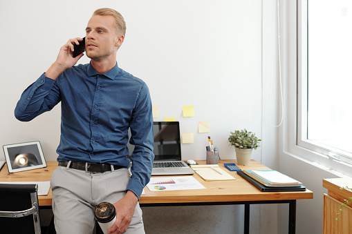 Attractive young entreprneur with disposable cup of coffee leaning on office desk and talking on phone with business partner or client