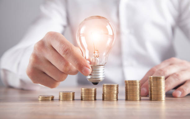 istock Man holding light bulb. Stack of coins on the desk. Saving energy and money 1314481650