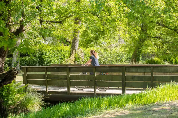 Photo of mature woman crossing the river by bicycle on a small wooden bridge