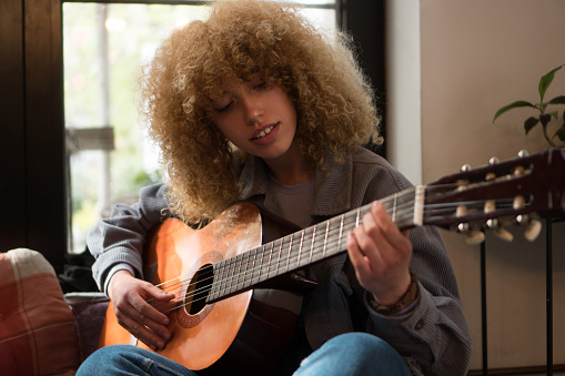A beautiful teenage girl with a curly hair is practicing acoustic guitar inside the house.
