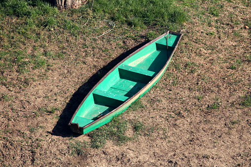 Elongated light green with dark brown edge wooden river boat left on grass and dry soil covered river bank tied with strong metal chain and cable to large tree on warm sunny winter day