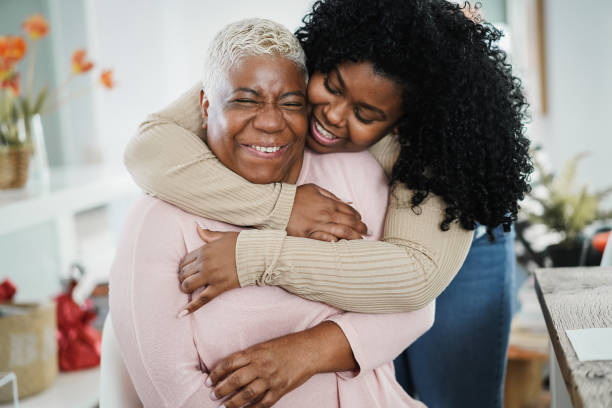 la figlia africana che abbraccia sua madre al chiuso a casa - focus principale sul viso della donna anziana - grandmother senior adult smiling women foto e immagini stock