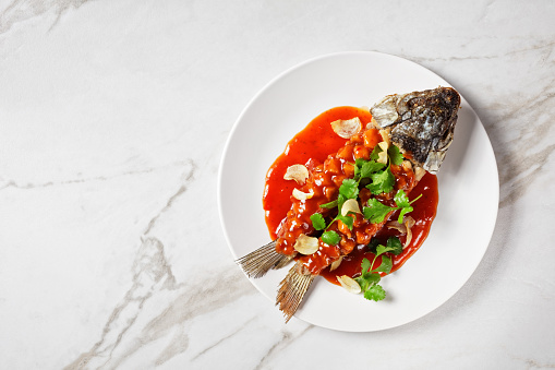 Carp squirrel or songshu yu under sweet and sour sauce and fresh cilantro served on a white plate on a white marble stone background, top view, copy space, close-up