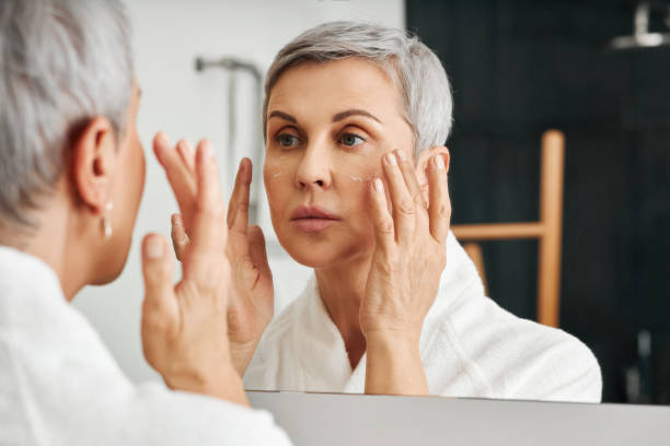 senior kvinna applicera fuktkräm i badrummet tittar på hennes reflektion - äldre kvinna hudvård bildbanksfoton och bilder