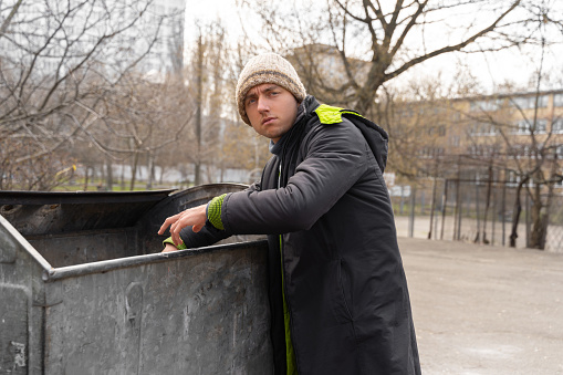 young homeless and unemployed man looking for food in dumpsters looking straight into the camera lens. poverty among homeless people forced to live on the street