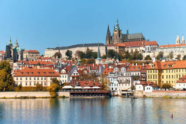 Photo of St. Vitus Cathedral and Prague Castle with orange roofs of historic buildings of Mala Strana reflected in river water on a bright Summer day in Prague, Czech Republic