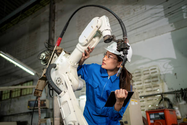 la mujer técnico revisa la máquina robótica del brazo en la fábrica. trabajador con casco de seguridad, gafas y uniforme. mantenimiento preventivo para evitar averías. concepto de servicio y reparación - manejar una máquina fotos fotografías e imágenes de stock