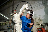 Technician woman check the robotic arm machine in the factory. Worker wearing safety helmet, glasses and uniform. Preventive maintenance to prevent breakdown. Service and repair concept
