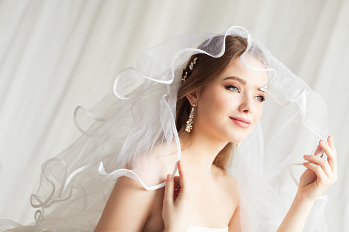 Bride in Veil. Wedding Make up and Bridal Hairstyle Fashion Portrait. Romantic happy Smiling Bride Looking Away at Window and Dreaming. Beauty Wedding Woman Face Close up over White Background