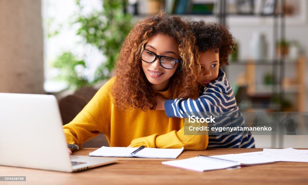 Young concentrated afro american woman mother working online on Working with kids. Young focused woman mother wearing eyeglasses using laptop and thinking about work task while small boy son gently hugs her. Childcare concept Working Mother Stock Photo