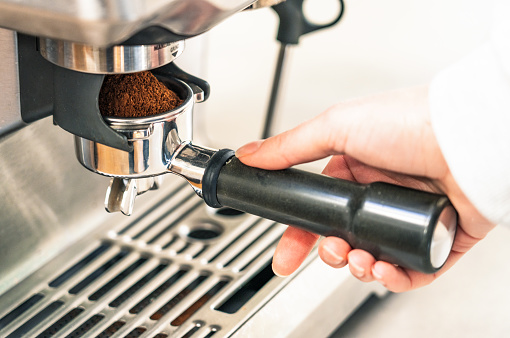 A barista holding a portafilter in the grinder holder of a coffee machine, with freshly ground coffee piled up in the portafilter.