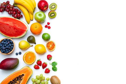 Overhead view of multicolored fresh ripe organic fruits shot on white background. The composition is at the left of an horizontal frame leaving useful copy space for text and/or logo at the right and includes mango, orange, strawberry, blueberry, kiwi, peach, grape, watermelon, banana, papaya, apple, pear, fig, lime and lemon. High resolution 42Mp studio digital capture taken with SONY A7rII and Zeiss Batis 40mm F2.0 CF lens