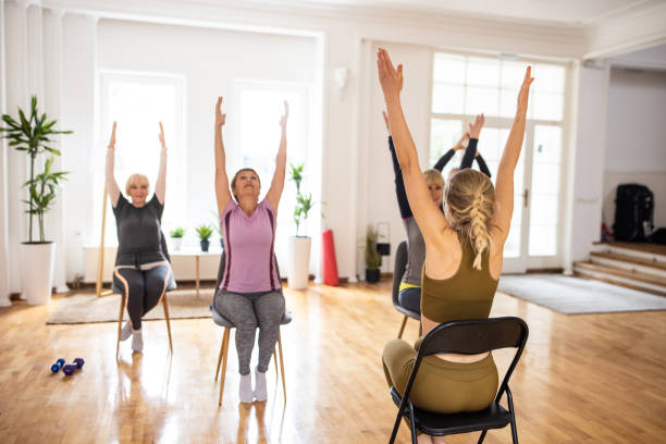 Female yoga instructor having yoga class with senior people on chairs Female yoga instructor having yoga class with senior people on chairs in yoga studio chair stock pictures, royalty-free photos & images