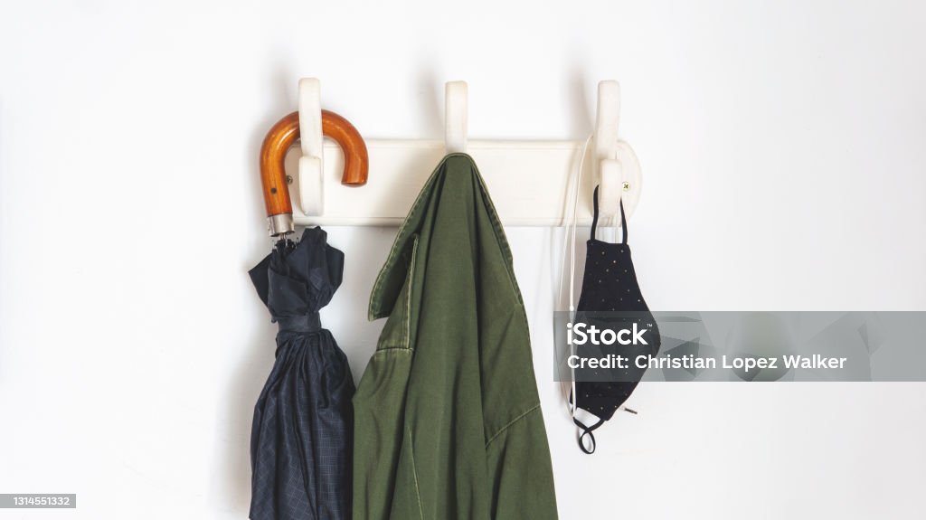 new normal things you need if you are going outside in the new normal because of covid-19. Hanging from a coat rack an umbrella, a coat, face masks and headphones. Over a white wall. Taken inddor with natural daylight Coat Hook Stock Photo