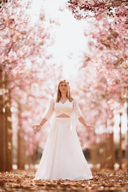 linda mulher loira em um vestido branco rezando com os braços abertos em pé em um bosque de primavera com árvores rosa florescendo - fairy forest women magic - fotografias e filmes do acervo