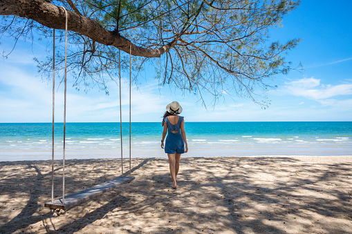 Asian woman wearing hat walking on the beach and wooden swing hanging from tree in tropical sea. Summer and holidays concept