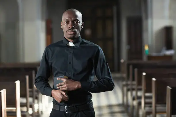 Portrait of African priest in black suit holding bible book and looking at camera standing in church