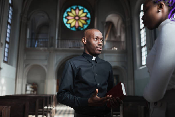 Priest talking to woman African young priest talking to woman and discussing the parts of Bible standing in the church place of worship stock pictures, royalty-free photos & images