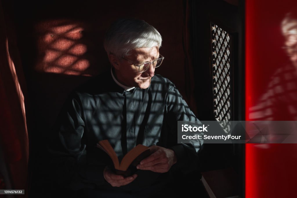 Priest in the confessional Senior priest sitting with the Bible in confessional and listening to confession Confession Booth Stock Photo