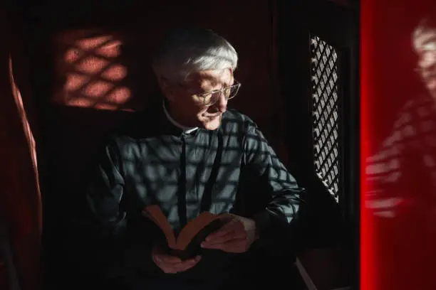 Senior priest sitting with the Bible in confessional and listening to confession