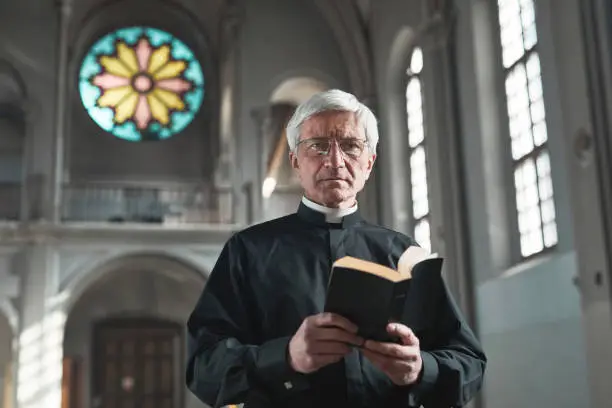 Portrait of senior priest holding the Bible and looking at camera while standing in the church