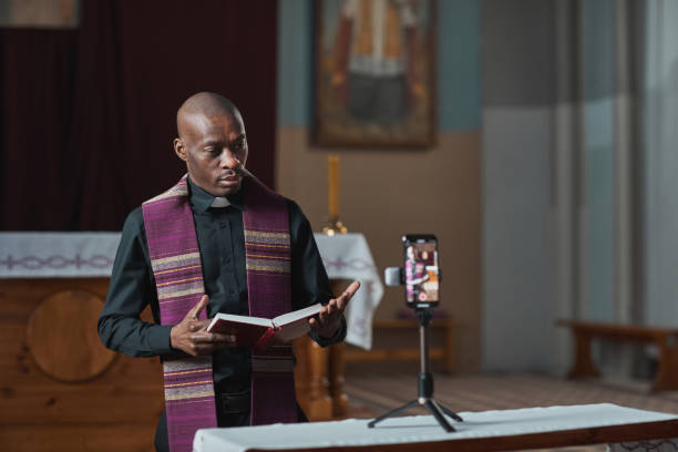 Priest performing online in the church African priest holding Bible and performing during live broadcast on mobile phone online clergy stock pictures, royalty-free photos & images