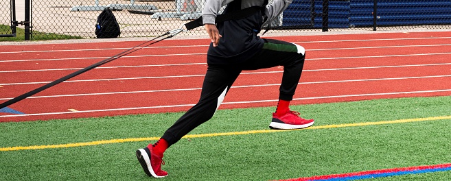 A male high school runner is dragging a sled with weight across a green turf field during strength and agility practice.