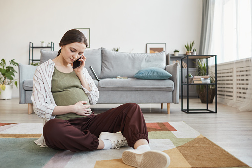 Pregnant woman sitting on the floor and talking on mobile phone in the living room