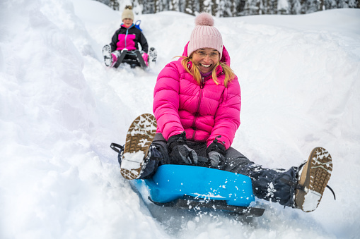 Smiling woman and girl enjoying tobogganing on snowy hill during winter.
