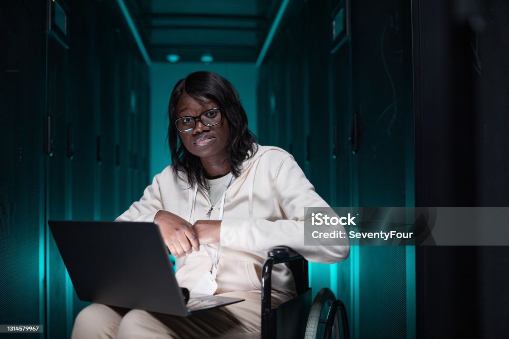 Accessible Workplace Portrait of disabled African-American woman using laptop and looking at camera while working in server room, accessible job opportunity, copy space Data Scientist Stock Photo