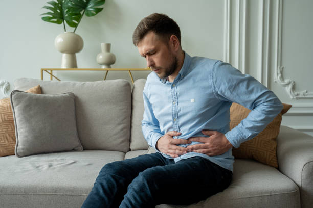 A man suffers from abdominal pain while sitting at home on the couch. Beautiful young man suffering from abdominal pain sitting on the couch at home A man suffers from abdominal pain while sitting at home on the couch. Beautiful young man suffering from abdominal pain sitting on the couch at home. stomachache stock pictures, royalty-free photos & images