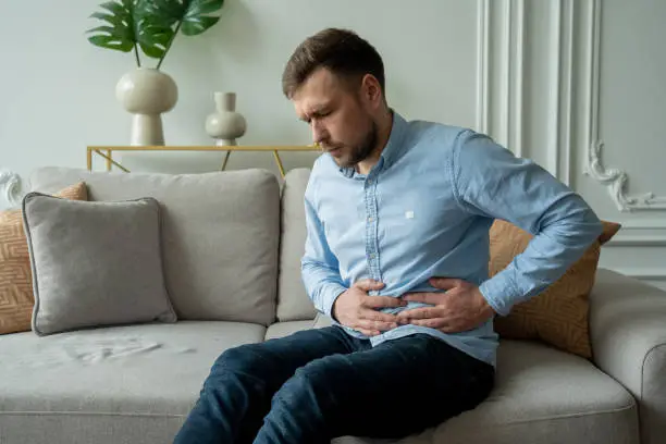 Photo of A man suffers from abdominal pain while sitting at home on the couch. Beautiful young man suffering from abdominal pain sitting on the couch at home