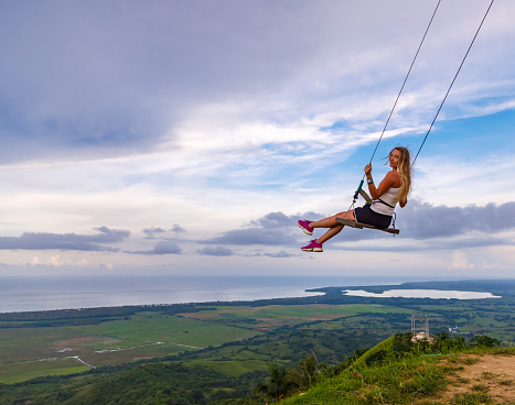 A young girl, blonde, swinging on a swing on a mountain slope in summer. Swing high in the mountains above the valley. Dominican Republic. Setting sun