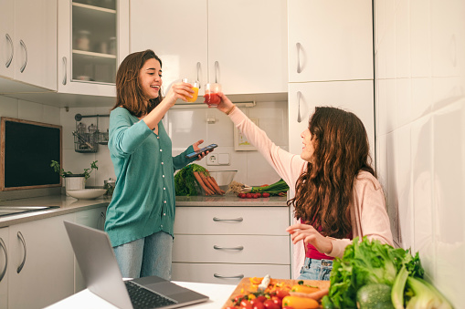Two cute sisters, in funny mood, toasting with natural juices, while preparing vegetarian food, in quarantine time