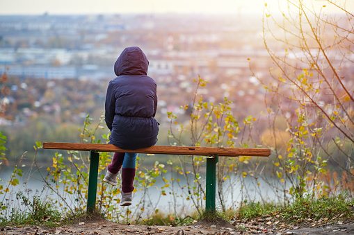 Nizhny Novgorod - 10.09.2019 - Young woman sitting on bench in autumn city park and looking at cityscape, back view. Self-reflection of girl, thinking about unrequited love for Valentines Day. Introspection concept.