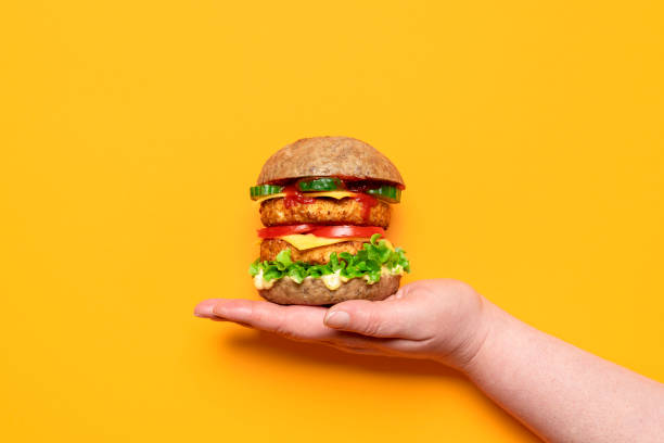Veggie burger top view, isolated on orange background. Hand holding soy burger Woman's hand holding a homemade vegan burger on an orange background. Veggie burger with soy patties, vegan cashew cheese, remoulade sauce and vegetables meat substitute stock pictures, royalty-free photos & images